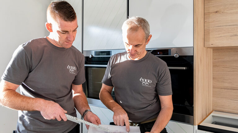 Deux hommes en t-shirts gris mesurent un plan devant des armoires de cuisine blanches et en bois clair.